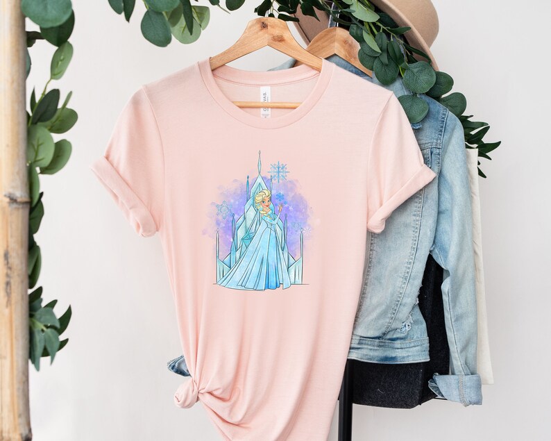 K&ouml;nnte beinhalten: Ein rosa T-Shirt mit einer Aquarell-Illustration von Elsa aus Frozen, die vor ihrem Eisschloss steht.