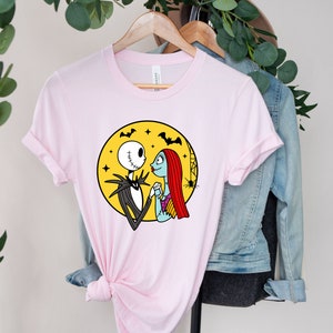 Pu&ograve; includere: Maglietta rosa con una grafica di Jack Skellington e Sally di The Nightmare Before Christmas. La grafica li mostra mano nella mano davanti a una luna gialla con pipistrelli e stelle.