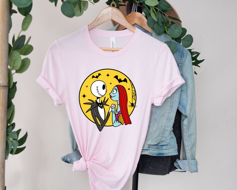 Pu&ograve; includere: Maglietta rosa con una grafica di Jack Skellington e Sally di The Nightmare Before Christmas. La grafica li mostra mano nella mano davanti a una luna gialla con pipistrelli e stelle.