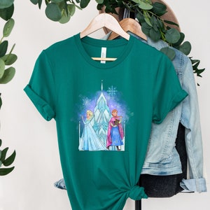K&ouml;nnte beinhalten: Ein gr&uuml;nes T-Shirt mit einem Aufdruck von Elsa und Anna aus Frozen, die vor einem Eis-Schloss stehen. Der Aufdruck ist im Aquarell-Stil gehalten.