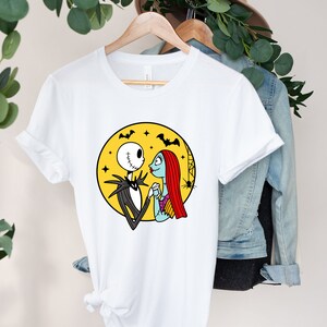 Pu&ograve; includere: Maglietta bianca con una grafica di Jack Skellington e Sally di The Nightmare Before Christmas. La grafica li mostra mano nella mano e si guardano di fronte a una luna gialla con pipistrelli che volano intorno.