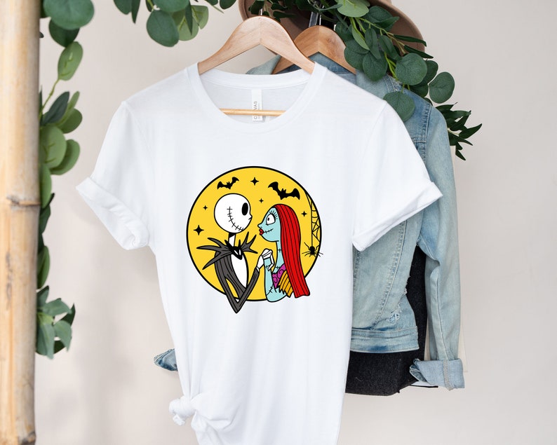 Pu&ograve; includere: Maglietta bianca con una grafica di Jack Skellington e Sally di The Nightmare Before Christmas. La grafica li mostra mano nella mano e si guardano di fronte a una luna gialla con pipistrelli che volano intorno.
