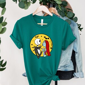 Pu&ograve; includere: Una maglietta verde turchese con una stampa grafica di Jack Skellington e Sally del film L'incubo prima di Natale. La grafica li mostra in piedi insieme davanti a una luna gialla con pipistrelli che volano intorno.