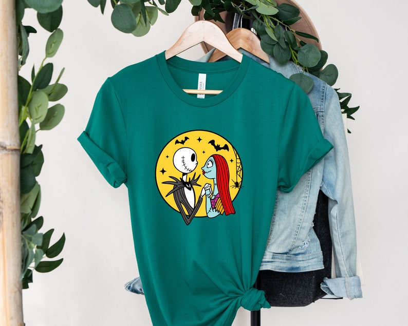 Pu&ograve; includere: Una maglietta verde turchese con una stampa grafica di Jack Skellington e Sally del film L'incubo prima di Natale. La grafica li mostra in piedi insieme davanti a una luna gialla con pipistrelli che volano intorno.