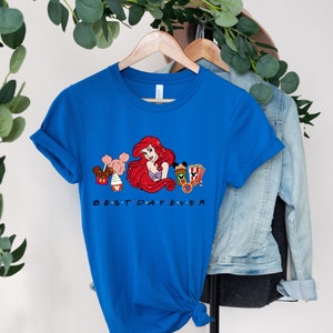 Op de afbeelding: Een koninklijk blauw T-shirt met een afbeelding van Ariel uit De Kleine Zeemeermin, Mickey Mouse-oren en Disney-park snacks. De tekst "BEST DAY EVER" is onder de afbeelding gedrukt.