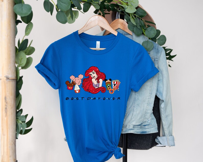 Op de afbeelding: Een koninklijk blauw T-shirt met een afbeelding van Ariel uit De Kleine Zeemeermin, Mickey Mouse-oren en Disney-park snacks. De tekst "BEST DAY EVER" is onder de afbeelding gedrukt.