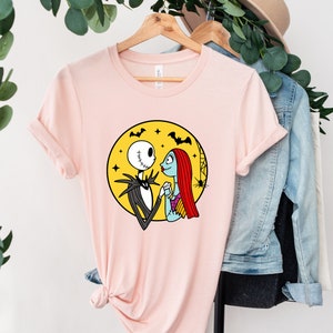 Pu&ograve; includere: Una maglietta rosa con una grafica di Jack Skellington e Sally di The Nightmare Before Christmas. La grafica li mostra mano nella mano davanti a una luna gialla con pipistrelli che volano intorno.