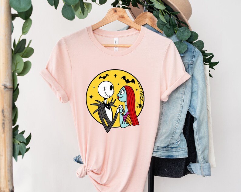 Pu&ograve; includere: Una maglietta rosa con una grafica di Jack Skellington e Sally di The Nightmare Before Christmas. La grafica li mostra mano nella mano davanti a una luna gialla con pipistrelli che volano intorno.