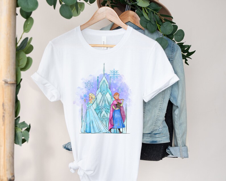 K&ouml;nnte beinhalten: Wei&szlig;es T-Shirt mit einer Aquarell-Illustration von Elsa und Anna aus Frozen, die vor dem Eisschloss stehen. Die Illustration ist in Blaut&ouml;nen, Lila und Rosa gehalten.