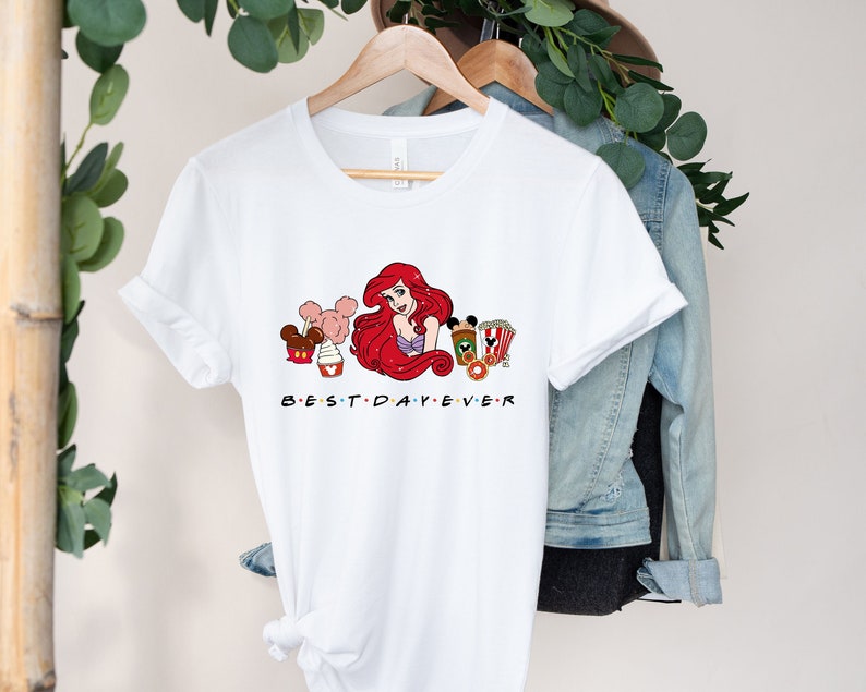 Op de afbeelding: Wit T-shirt met een afbeelding van Ariel uit De Kleine Zeemeermin, Mickey Mouse-oren en andere Disney-snacks. De tekst "BEST DAY EVER" is onder de afbeelding gedrukt.