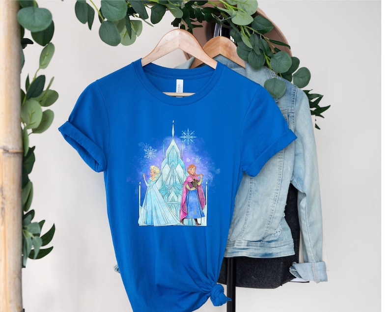K&ouml;nnte beinhalten: Ein k&ouml;nigsblaues T-Shirt mit einem Aufdruck von Elsa und Anna aus Frozen, die vor ihrem Eisschloss stehen. Der Aufdruck ist im Aquarellstil gehalten und in einem hellblauen, violetten und rosafarbenen Farbschema gehalten.