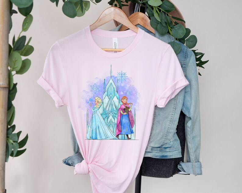 K&ouml;nnte beinhalten: Pinkes T-Shirt mit einer Aquarell-Illustration von Elsa und Anna aus Frozen, die vor ihrem Eisschloss stehen. Das Schloss ist blau und wei&szlig;, und die Figuren tragen ihre charakteristischen Kleider.