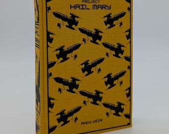 Op de afbeelding: Een hardcover boek getiteld "Project Hail Mary" van Andy Weir. De omslag is geel met een herhalend patroon van zwarte ruimteschip illustraties en kleine figuren. De rug van het boek bevat ook de titel.