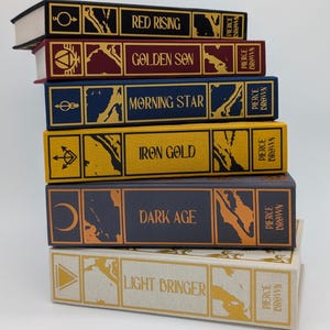 May include: A stack of six hardcover books with gold lettering and decorative designs. The titles include "Red Rising," "Golden Son," "Morning Star," "Iron Gold," "Dark Age," and "Light Bringer." Each book has a different color scheme, with the author's name, Pierce Brown, printed on the spine.