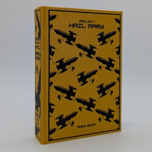 Puede incluir: Un libro de tapa dura titulado Project Hail Mary de Andy Weir con una cubierta de tela color amarillo mostaza. La parte frontal presenta un patrón negro repetitivo de naves espaciales y astronautas. El lomo incluye el título y el autor.