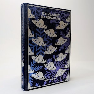 May include: A hardcover book titled "Ice Planet Barbarians" by Ruby Dixon. The cover is dark blue with silver UFO illustrations and snowflakes. The book's spine also features the title.