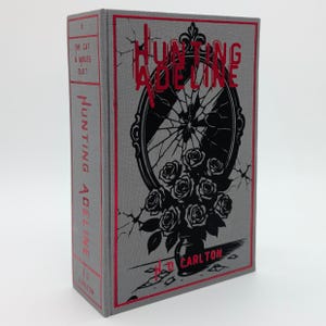 May include: A hardcover book titled "Hunting Adeline" with a gray cover and red accents. The cover features a black and white illustration of a broken mirror and roses. The author's name, H.D. Carlton, is printed at the bottom.