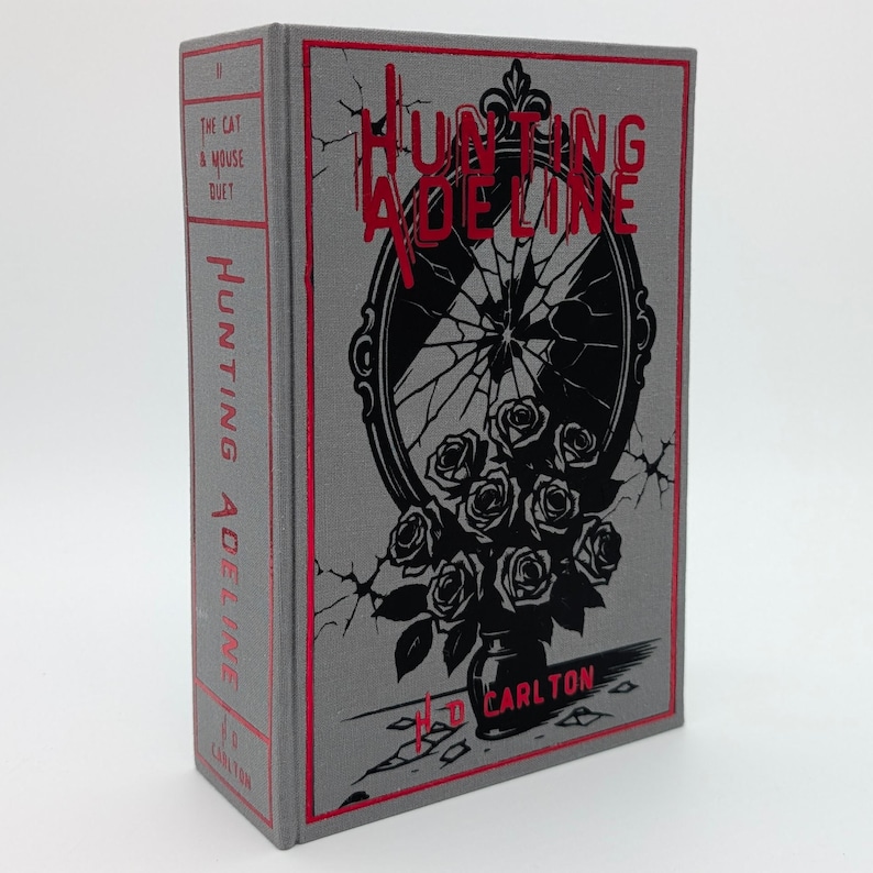 May include: A hardcover book titled "Hunting Adeline" with a gray cover and red accents. The cover features a black and white illustration of a broken mirror and roses. The author's name, H.D. Carlton, is printed at the bottom.