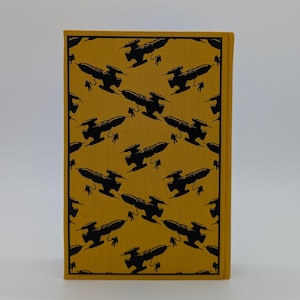 May include: A rectangular book with a mustard yellow fabric cover featuring a repeating black graphic pattern of spaceships and small figures of astronauts floating in space, arranged in a diagonal grid across the entire front surface.