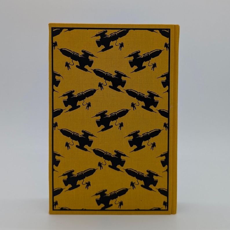 May include: A rectangular book with a mustard yellow fabric cover featuring a repeating black graphic pattern of spaceships and small figures of astronauts floating in space, arranged in a diagonal grid across the entire front surface.