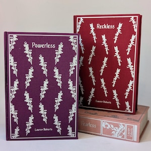 Può includere: Una pila di libri con motivi floreali. Il libro in alto è viola scuro con il titolo "Powerless" e il nome dell'autore. Il libro successivo è rosso con il titolo "Reckless". Il libro in basso è rosa con il titolo "Fearless".