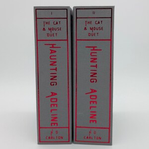 May include: Two hardcover books, "The Cat & Mouse Duet" series, are displayed. The spines are gray with red text that reads "Haunting Adeline" and "Hunting Adeline". The publisher's name, "H.D. Carlton", is at the bottom.
