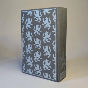 May include: A hardcover book with a gray cover featuring a repeating light blue lion pattern. The spine has a light blue lion design and the author's name, George R.R. Martin, is printed at the bottom. The book is a fantasy novel.