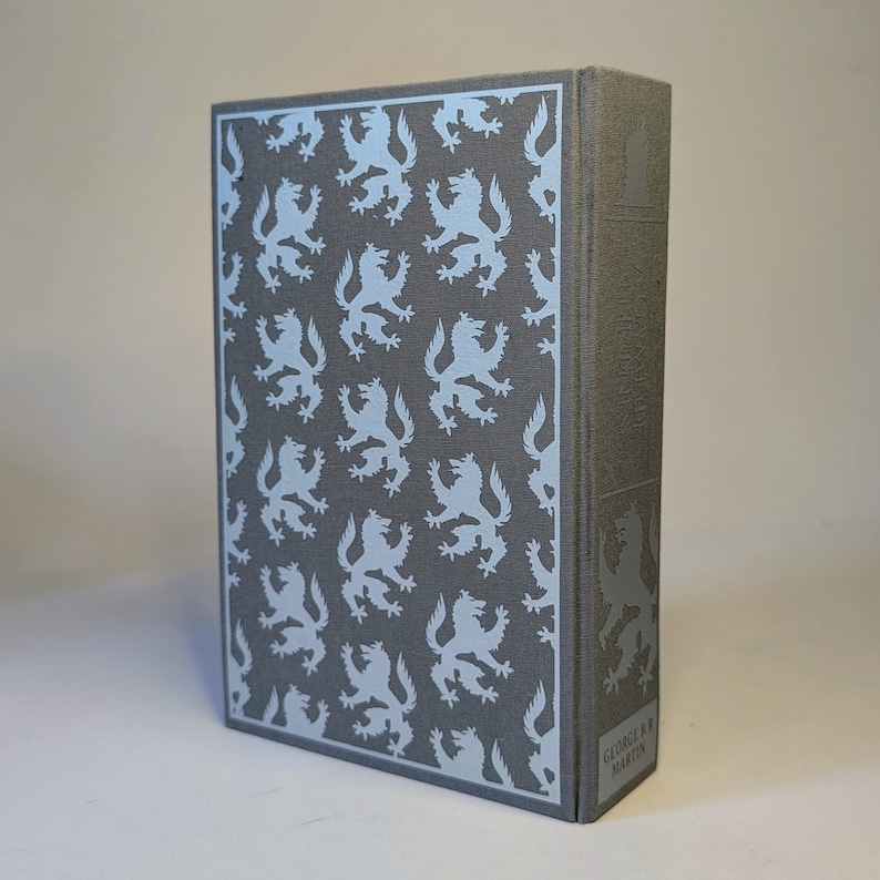 May include: A hardcover book with a gray cover featuring a repeating light blue lion pattern. The spine has a light blue lion design and the author's name, George R.R. Martin, is printed at the bottom. The book is a fantasy novel.