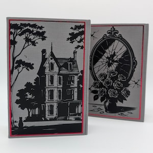 May include: Two hardback books with gray covers and red trim. One book features a black illustration of a house and a tree. The other book has a black illustration of a broken mirror and roses in a vase. The books are standing upright.