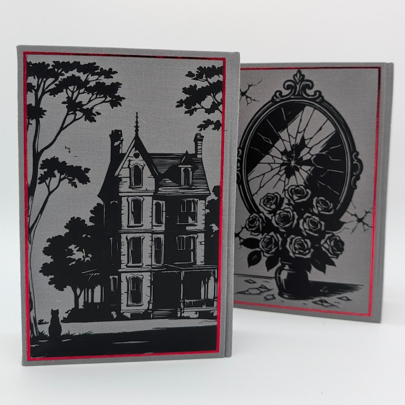May include: Two hardback books with gray covers and red trim. One book features a black illustration of a house and a tree. The other book has a black illustration of a broken mirror and roses in a vase. The books are standing upright.