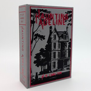 May include: A hardcover book titled "Haunting Adeline" with a gray cover and red accents. The cover features a black illustration of a Victorian house, trees, and the author's name, M.D. Carlton. The spine also displays the title.