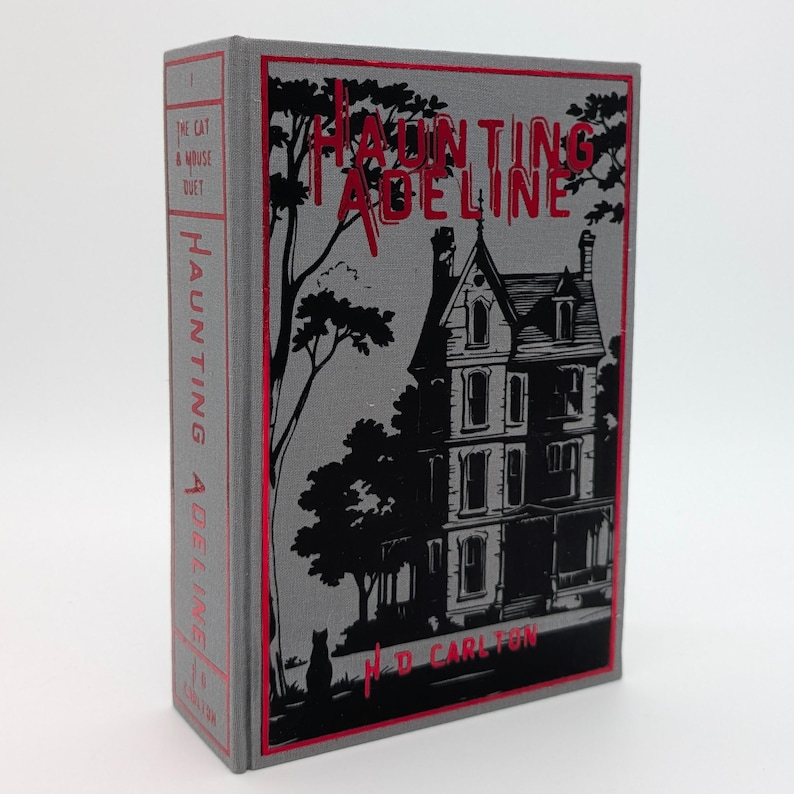 May include: A hardcover book titled "Haunting Adeline" with a gray cover and red accents. The cover features a black illustration of a Victorian house, trees, and the author's name, M.D. Carlton. The spine also displays the title.