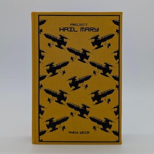 May include: A mustard yellow hardcover book titled Project Hail Mary by Andy Weir. The cover features a repeating black graphic pattern of spaceships and astronauts tethered to them, arranged in a diagonal grid across the entire front surface.