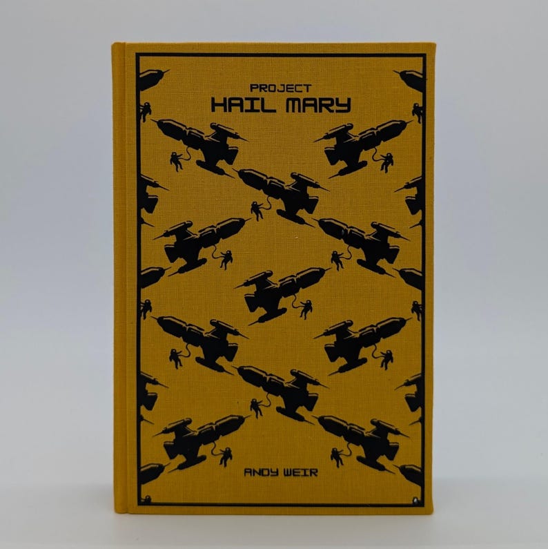 May include: A mustard yellow hardcover book titled Project Hail Mary by Andy Weir. The cover features a repeating black graphic pattern of spaceships and astronauts tethered to them, arranged in a diagonal grid across the entire front surface.