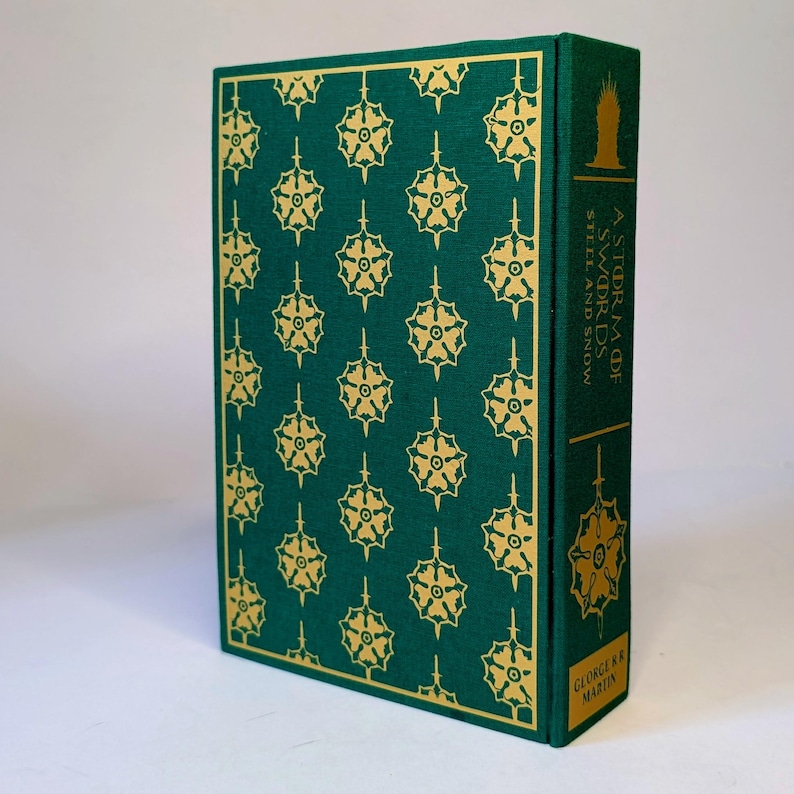 May include: A hardcover book with a green cover featuring a repeating gold floral pattern. The spine displays the title "A Storm of Swords" and the author's name, George R.R. Martin. The book's design is detailed and decorative.