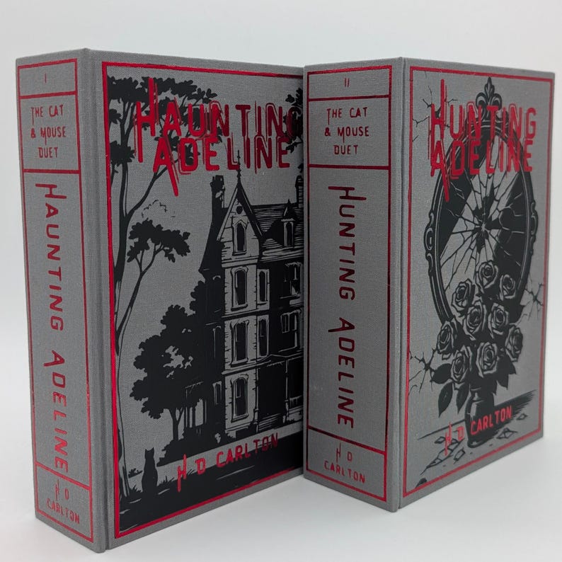 May include: Two hardcover books titled "Hunting Adeline" with matching designs. The covers are gray with red accents and feature illustrations of a house, a mirror with roses, and the title in a stylized font. The books are part of "The Cat & Mouse Duet" series.