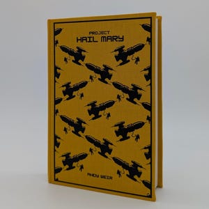 May include: A hardcover book titled Project Hail Mary by Andy Weir with a mustard yellow fabric cover. The front features a repeating black graphic pattern of spaceships and astronauts, enclosed within a thin black rectangular border.