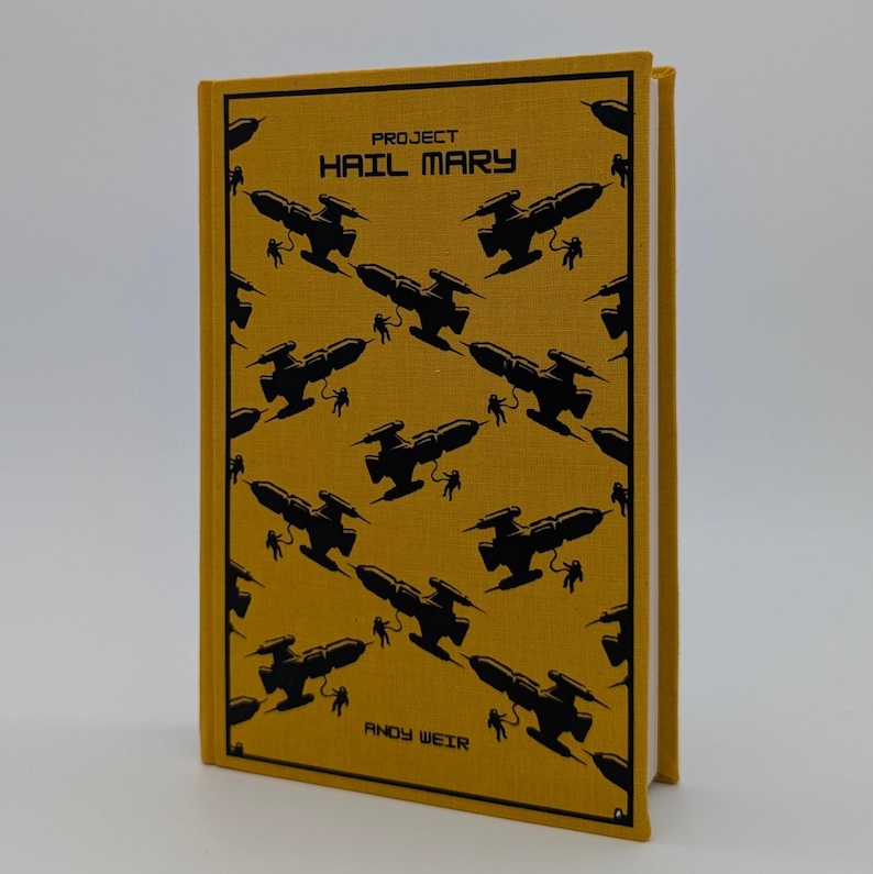 May include: A hardcover book titled Project Hail Mary by Andy Weir with a mustard yellow fabric cover. The front features a repeating black graphic pattern of spaceships and astronauts, enclosed within a thin black rectangular border.