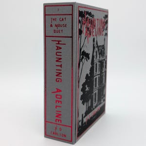 May include: A hardcover book titled "Haunting Adeline" with a gray cover and red lettering. The spine reads "The Cat & Mouse Duet" and "Haunting Adeline". The cover features a black and white illustration of a house and trees.