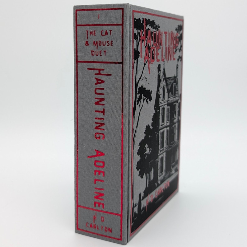 May include: A hardcover book titled "Haunting Adeline" with a gray cover and red lettering. The spine reads "The Cat & Mouse Duet" and "Haunting Adeline". The cover features a black and white illustration of a house and trees.