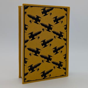 May include: A hardcover book with a mustard yellow fabric cover featuring a repeating black pattern of silhouettes depicting spaceships and astronauts floating in space. The book is standing upright, showing the front cover and the white page edges.