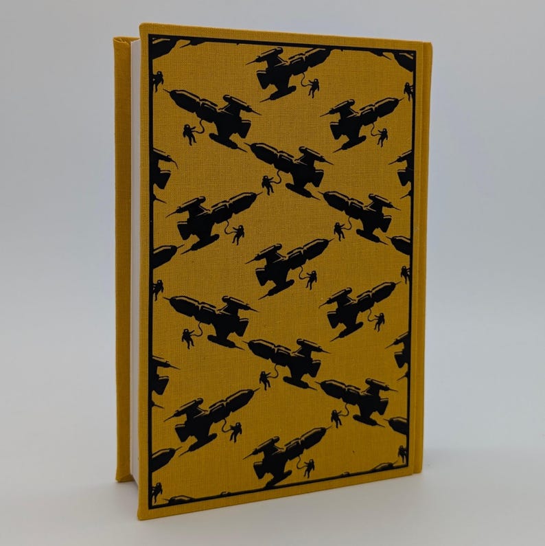 May include: A hardcover book with a mustard yellow fabric cover featuring a repeating black pattern of silhouettes depicting spaceships and astronauts floating in space. The book is standing upright, showing the front cover and the white page edges.