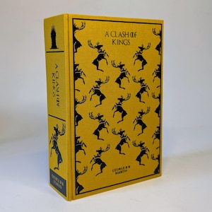 May include: A hardcover book titled "A Clash of Kings" by George R.R. Martin. The book has a yellow cover with a repeating pattern of black stags wearing crowns. The spine also features the title and author's name.