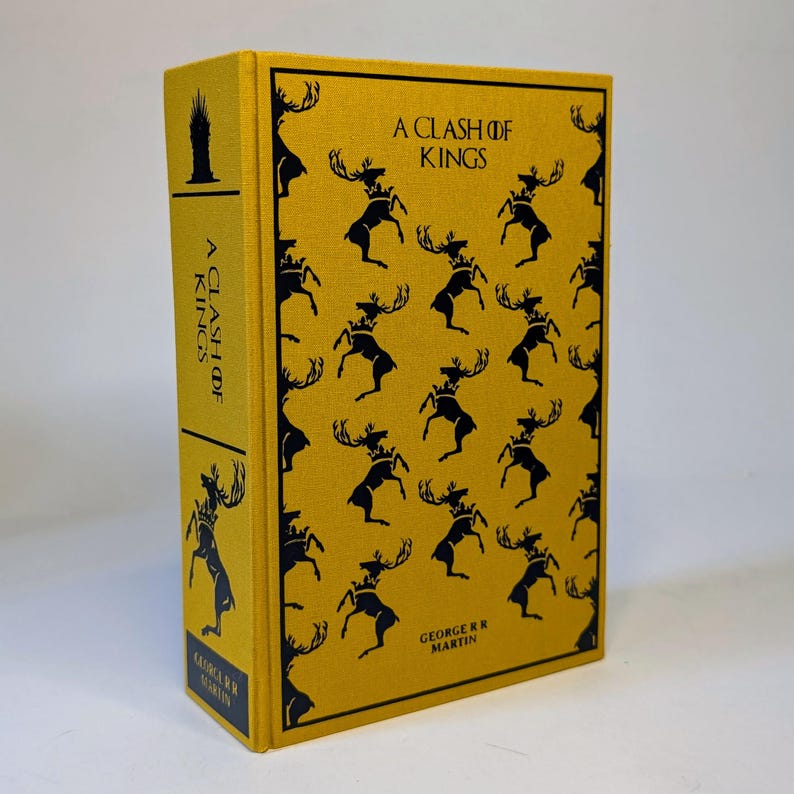 May include: A hardcover book titled "A Clash of Kings" by George R.R. Martin. The book has a yellow cover with a repeating pattern of black stags wearing crowns. The spine also features the title and author's name.