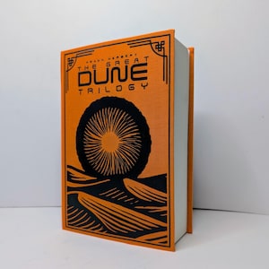 Puede incluir: Un libro de tapa dura titulado "The Great Dune Trilogy" de Frank Herbert. El libro tiene una cubierta naranja con elementos gráficos negros, incluyendo un diseño circular y líneas estilizadas. El lomo del libro también es naranja.