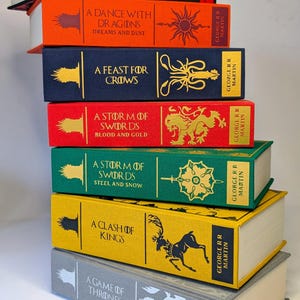 May include: A stack of seven hardcover books with colorful spines. The books feature titles like "A Dance with Dragons" and "A Game of Thrones" by George R.R. Martin. Each book has unique artwork and text on the spine, with white pages.