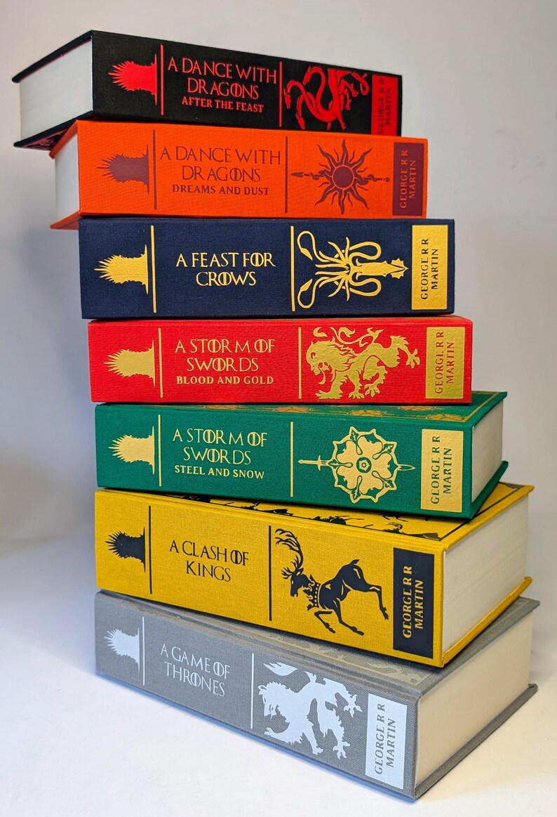 May include: A stack of seven hardcover books with colorful spines. The books feature titles like "A Dance with Dragons" and "A Game of Thrones" by George R.R. Martin. Each book has unique artwork and text on the spine, with white pages.