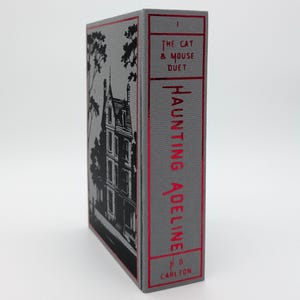 May include: A hardcover book titled "Haunting Adeline" with a gray cover and red lettering. The spine features the title and author's name, with the words "The Cat & Mouse Duet" at the top. The cover has a black illustration of a building.