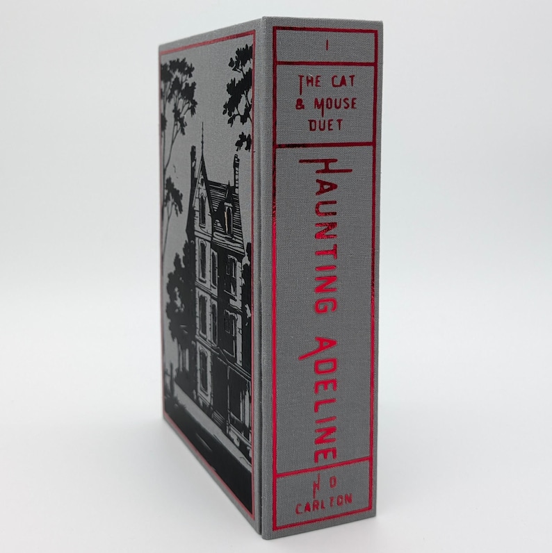 May include: A hardcover book titled "Haunting Adeline" with a gray cover and red lettering. The spine features the title and author's name, with the words "The Cat & Mouse Duet" at the top. The cover has a black illustration of a building.