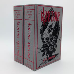 May include: Two hardcover books titled "Hunting Adeline" and "The Cat & Mouse Duet". The books have gray covers with red text and accents. The cover art features a black and white illustration of a broken mirror and roses.